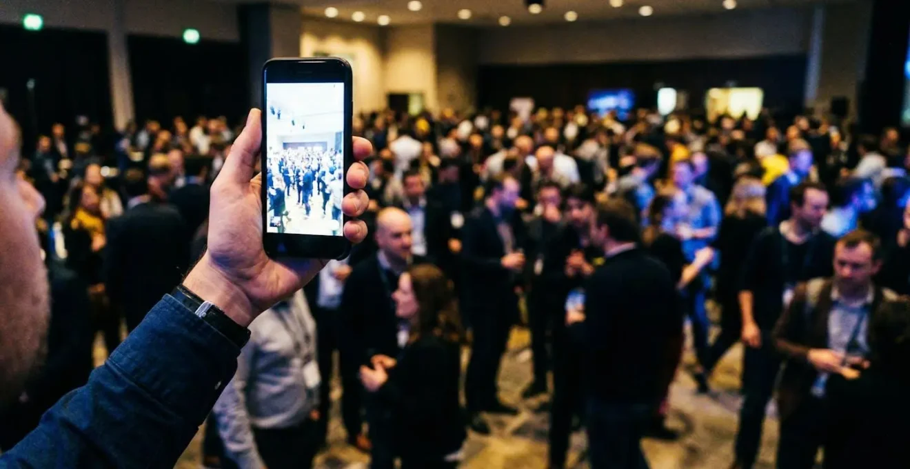 Ein Smartphone wird hochgehalten, um etwas zu filmen, unscharfer Bürohintergrund mit Personen